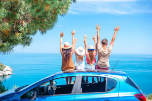 family sitting on top of blue car with hands up in the air with lake in background